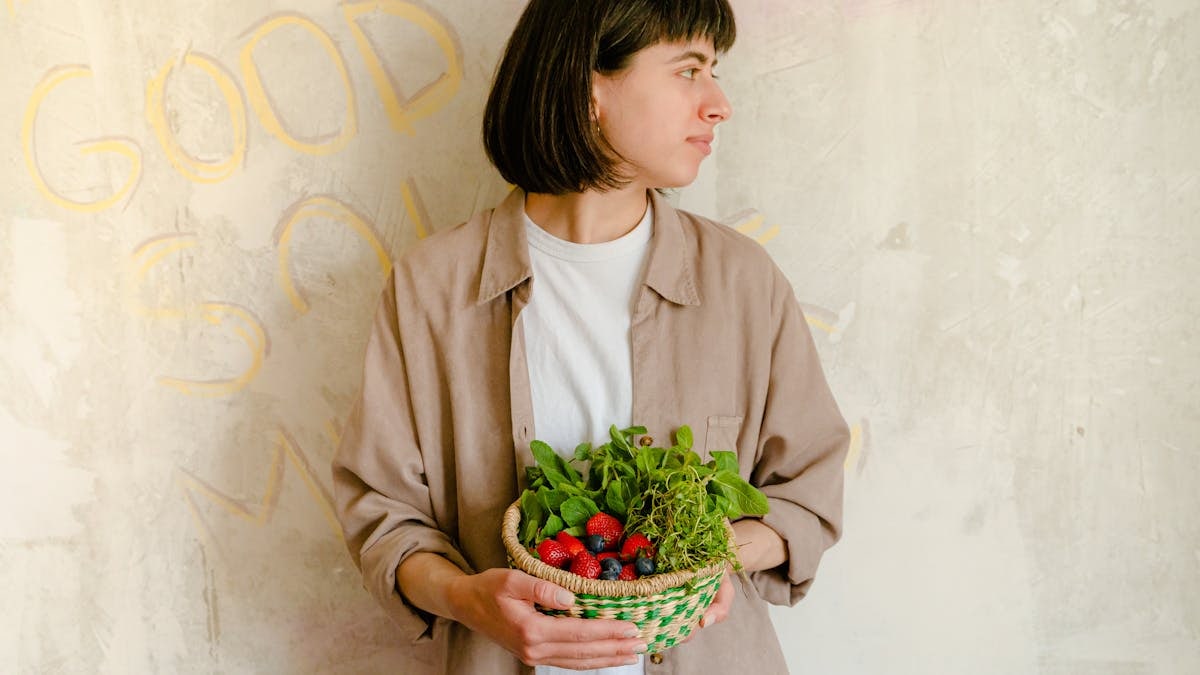 Woman standing with a basket of fresh greens and berries against a rustic wall background.